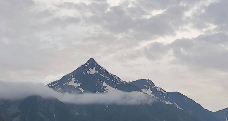 Snow-dusted mountain peak piercing through low clouds beneath a mottled grey sky.