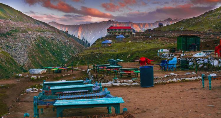 Outdoor mountainside café with colourful tables at dusk, dramatic peaks glowing in sunset light.