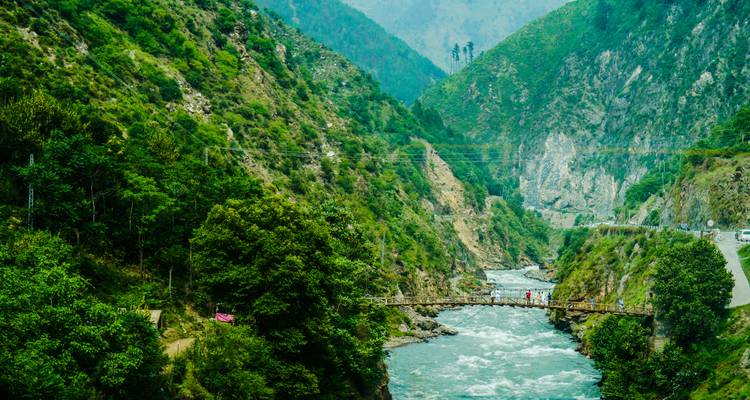 Narrow wooden bridge spans a fast-flowing river between steep green valley walls.