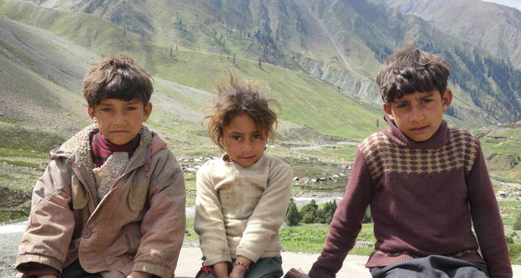 Three local children sit on a mountainside with valley terraces and peaks behind them.