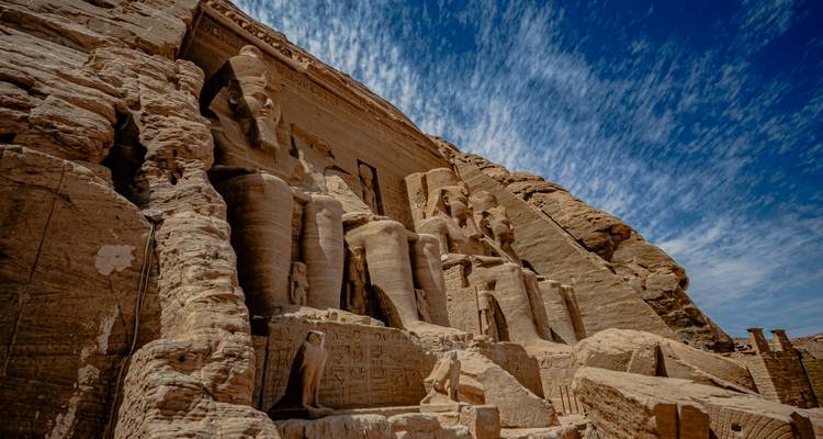 Vista angular amplia hacia arriba de las colosales estatuas sedentes talladas en el Templo de Abu Simbel bajo un cielo dramático con franjas.