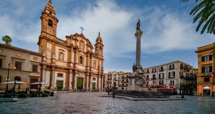 Plaza histórica en Palermo con fachada de iglesia barroca y estatua de columna alta al atardecer.