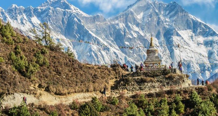 Wanderer, die auf einem Bergpfad an einem Stupa mit Gebetsfahnen vorbei gehen, unter hoch aufragenden schneebedeckten Himalaya-Gipfeln.
