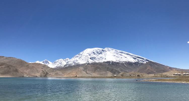Der schneebedeckte Muztagh Ata erhebt sich steil über einen klaren Alpensee und kahle Vorberge unter einem endlosen blauen Himmel.