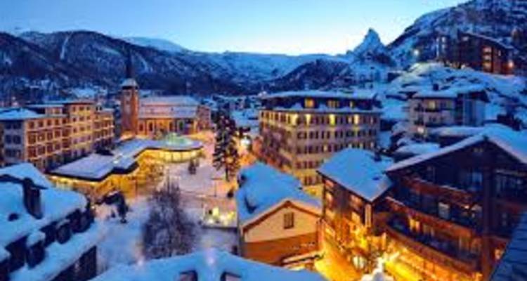 Verschneites Alpendorf in der Dämmerung mit warmbeleuchteten Straßen und Bergkulisse.