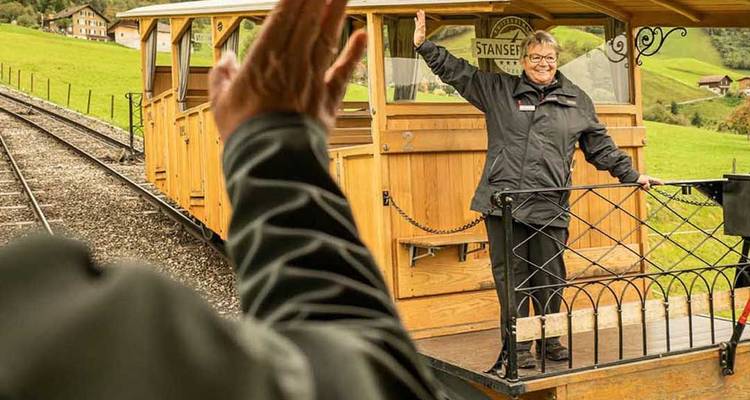 Glimlachend spoorwegpersoneel zwaait vanuit een openlucht houten wagon terwijl een andere persoon terugzwaait.