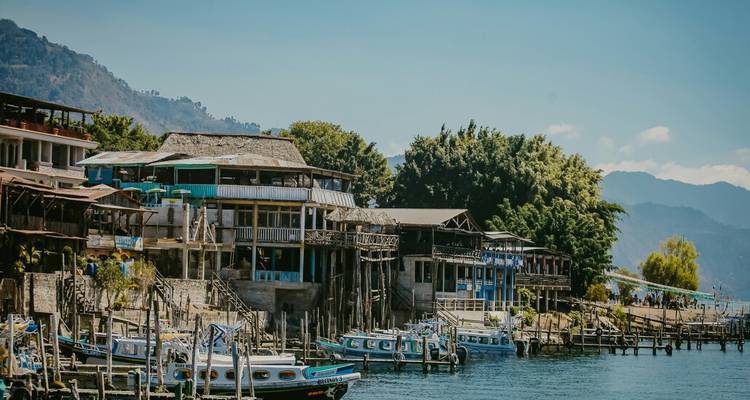 Muelles de madera junto al lago y edificios rústicos respaldados por colinas boscosas y montañas distantes bajo un cielo despejado.