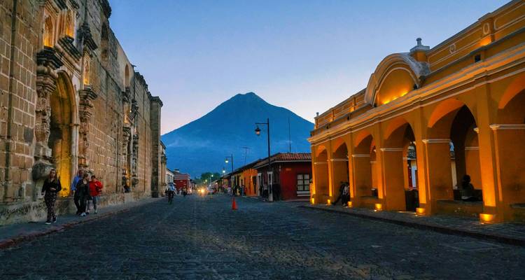 Calle empedrada al atardecer en Antigua con arco colonial iluminado y silueta de volcán al fondo.