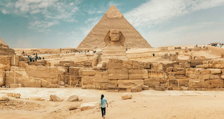 Un voyageur marche vers le Sphinx et la Grande Pyramide sous un ciel bleu éclatant.