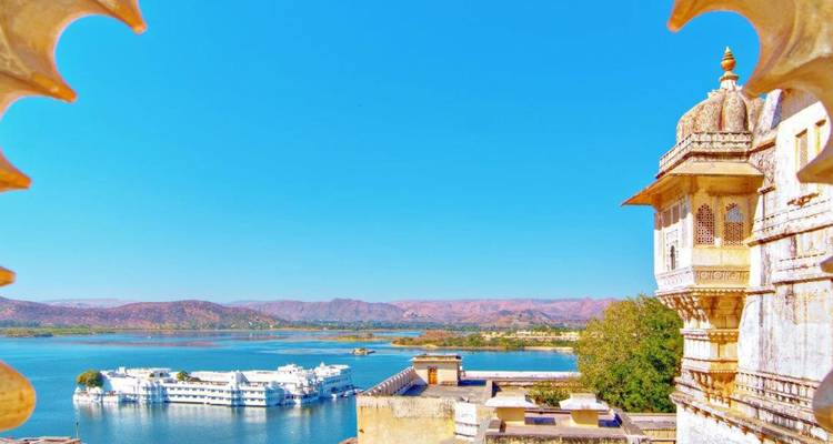 Panoramablick auf den Pichola-See mit dem weißen Seepalast, der auf blauem Wasser schwebt, eingerahmt von Palastbogendetails und rosa getönten Aravalli-Hügeln.