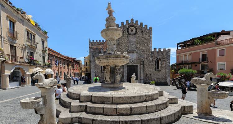 Fontaine historique en pierre entourée de sculptures devant le Duomo de Taormina sous un ciel bleu éclatant.