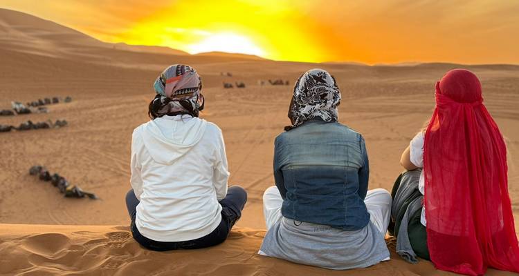 Drei Reisende in Kopftüchern sitzen auf einer Düne und beobachten den goldenen Sonnenaufgang über Kamelkarawanen in der Sahara.