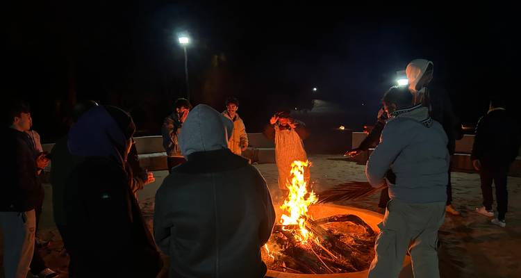 Reisende versammeln sich um ein loderndes Lagerfeuer unter dem Wüstennachthimmel, wärmen ihre Hände und unterhalten sich.