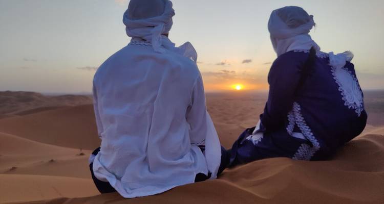 Zwei Reisende in traditioneller Wüstenkleidung sitzen auf einer Düne und beobachten den Sonnenuntergang über endlosen Sandwellen.