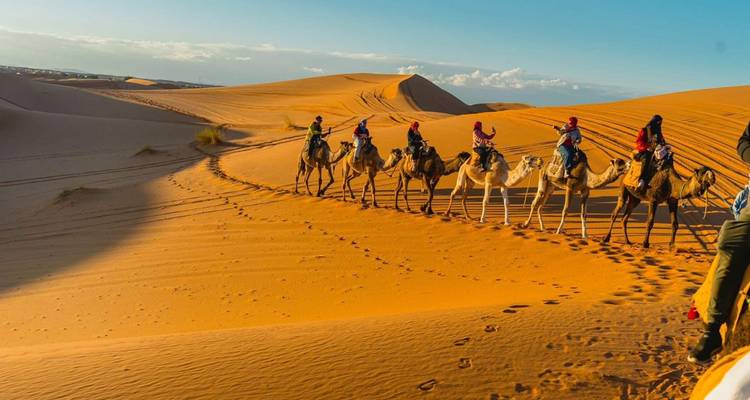 Kamelkarawane von Reitern schlängelt sich über goldene Sahara-Dünen unter klarem Himmel.