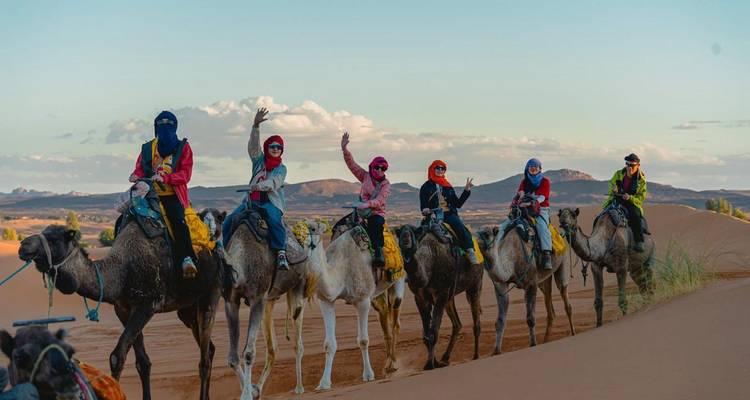Nahaufnahme von bunten Kamelreitern, die winken, während sie Wüstendünen im späten Nachmittagslicht durchqueren.