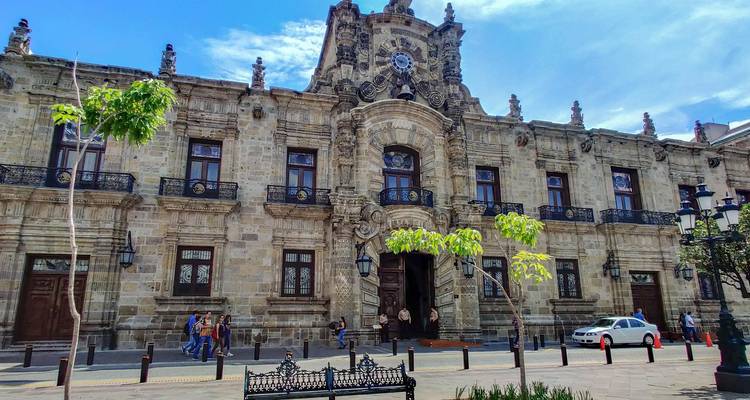Façade en pierre ornée du Palais du Gouvernement de Jalisco face à une petite place avec des piétons et un taxi.