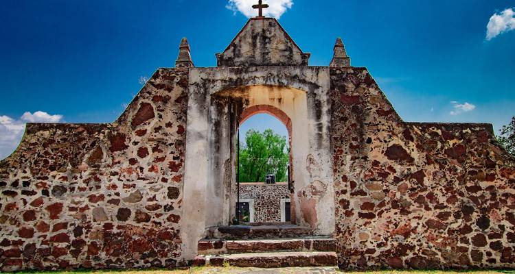 Une arche en pierre patinée avec une croix couronne un petit mur de style mission sous un ciel bleu profond.
