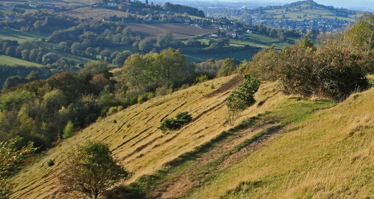 Le sentier de la colline herbeuse surplombe une campagne en patchwork et des villages lointains sous une lumière brumeuse.