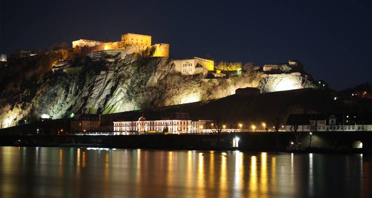 Beleuchtete Festung auf einem felsigen Hügel mit Blick auf einen ruhigen Fluss bei Nacht