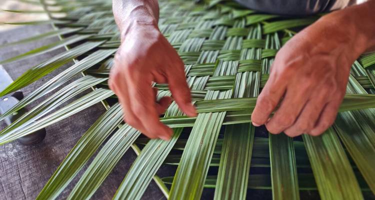 Close-up of hands weaving green palm fronds into a traditional mat on a wooden table.