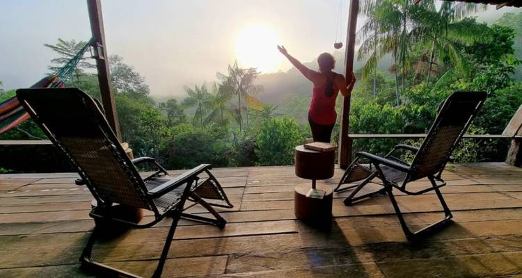 Person stretching arms toward the rising sun on a wooden jungle deck with lounge chairs and hammocks.