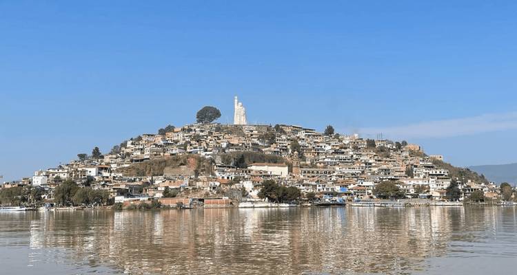 Ville à flanc de colline avec une grande statue au sommet se reflétant sur les eaux calmes du lac sous un ciel bleu dégagé.
