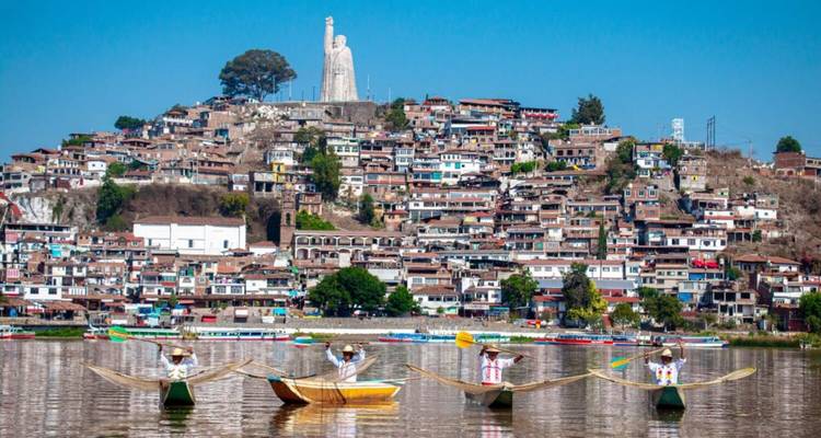 Des pêcheurs traditionnels dans des canoës décorés se produisent sur le lac avec la ville de Patzcuaro à flanc de colline et une statue en arrière-plan.