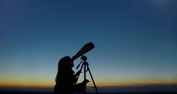 Silhouette einer Frau, die bei Einbruch der Dämmerung den Nachthimmel durch ein Teleskop beobachtet.
