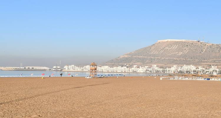 Amplia extensión arenosa de una playa vacía con la ciudad de Agadir y la fortaleza en la cima de la colina visibles a lo lejos.