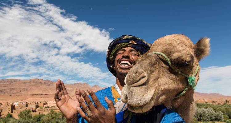 Homme souriant en tenue traditionnelle posant avec un chameau amical devant un village du désert et un ciel bleu.
