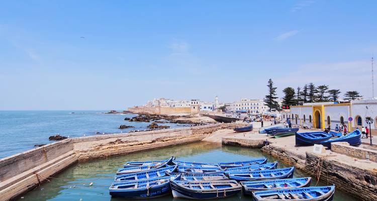 Le port d'Essaouira avec des rangées de bateaux en bois bleus amarrés dans une crique calme à côté des anciens remparts de la ville.