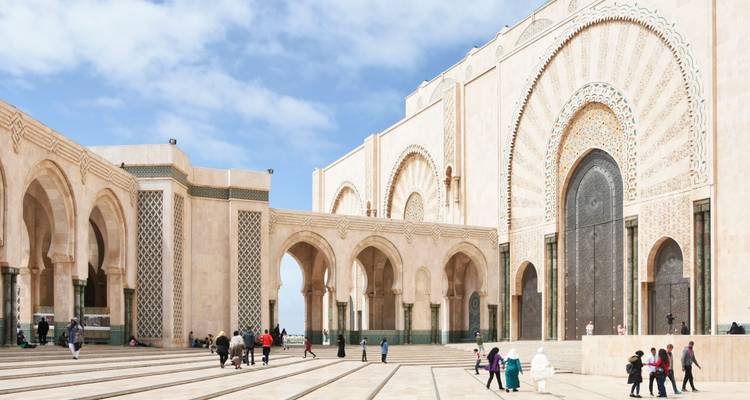 Der riesige Marmorhof der Hassan-II-Moschee in Casablanca mit kunstvollen Bögen und einer imposanten Verzierungstür, Besucher verstreut über den sonnigen Platz.