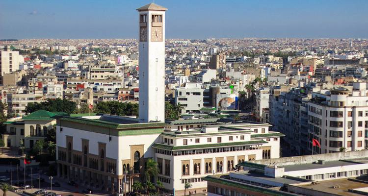 Eine Panoramablick auf die Stadtlandschaft von Casablanca, dominiert vom weissen Uhrenturm des Rathauses inmitten dicht bebauter städtischer Gebäude unter klarem blauen Himmel.
