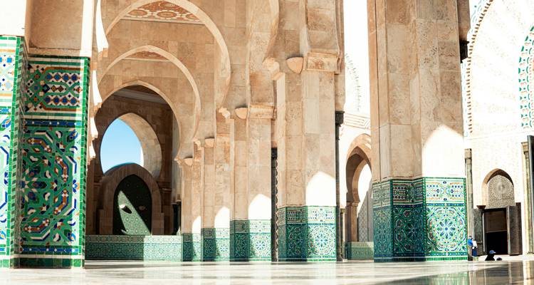 Sonnenlicht strömt durch verzierte Bögen und Säulen mit Mosaik-gekachelten Sockeln in der Hassan-II-Moschee und erzeugt geometrische Schatten auf dem polierten Boden.