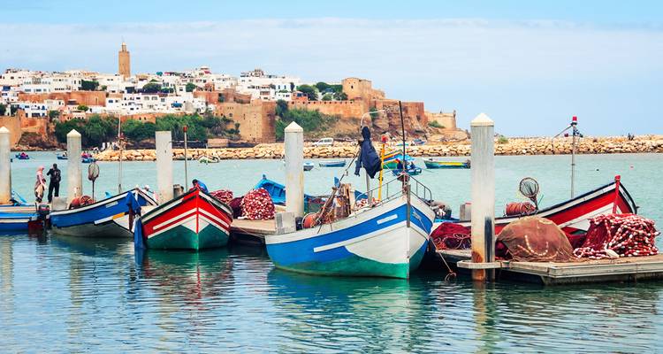 Botes de pesca pintados con colores brillantes se mecen en un muelle fluvial tranquilo con la Kasbah de los Udayas de Rabat visible en el acantilado detrás.