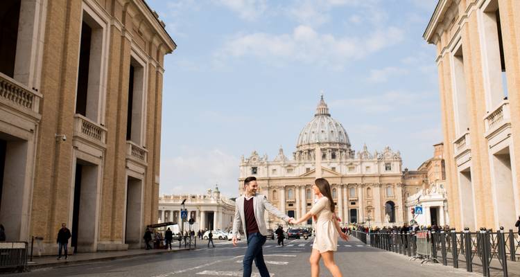 Un couple se tenant la main se promène vers la basilique Saint-Pierre le long d'une grande avenue bordée de bâtiments historiques.