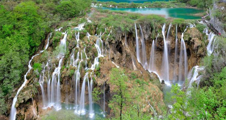 Panoramisch uitzicht op meerdere trapsgewijze watervallen die in smaragdgroene poelen stromen te midden van weelderig lentegebladerte.