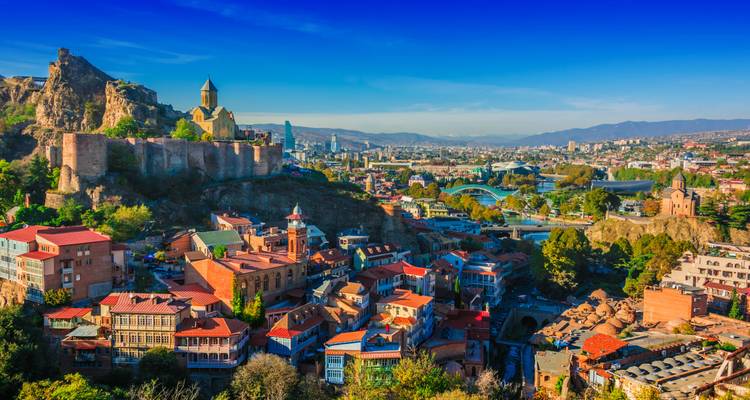 Panoramablick über Tiflis mit der Narikala-Festung und der rotbedachten Altstadt am Kura-Fluss unter einem klaren blauen Himmel.