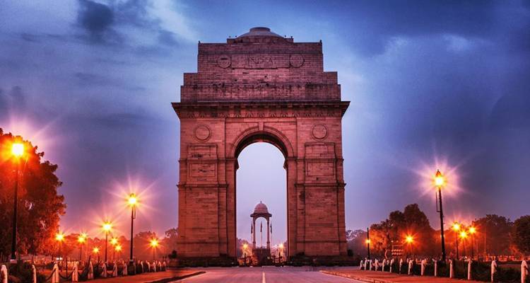 Das beleuchtete India Gate wölbt sich über eine leere Allee unter einem Dämmerhimmel.