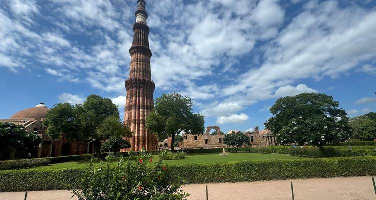 Der hohe rote Sandstein-Qutub-Minar erhebt sich über gepflegten Gärten unter einem wolkigen blauen Himmel.