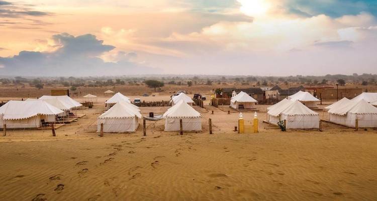 Weiße Leinwandzelte bilden ein Wüstenlager auf goldenem Sand unter einem farbenfrohen Abendhimmel.