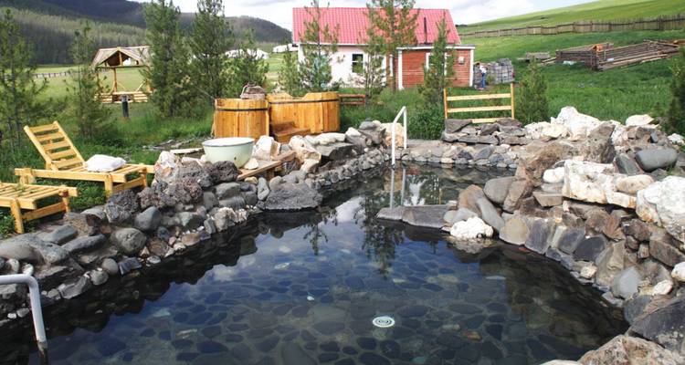 Piscine en pierre extérieure remplie d'eau minérale fumante située au milieu de collines herbeuses à côté de bacs en bois rustiques et de chaises longues.