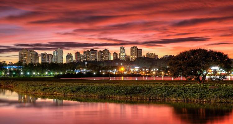 De levendige zonsondergang kleurt de skyline van Curitiba in rijke roodtinten, weerspiegeld in een meer op de voorgrond.
