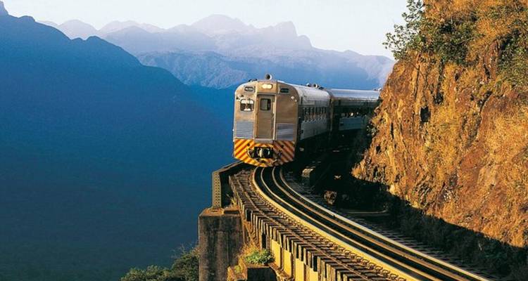 Serra Verde Express trein rijdt over een hoge bergbrug met blauwe bergkammen op de achtergrond.