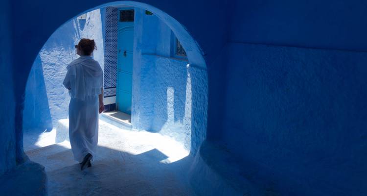 Mujer de blanco caminando por un callejón estrecho pintado de azul bañado en luz suave en Chefchaouen.