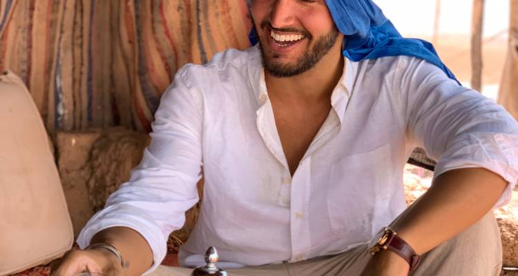 Hombre sonriente con camisa blanca y pañuelo azul en la cabeza sirviendo té tradicional dentro de una tienda estilo desierto.
