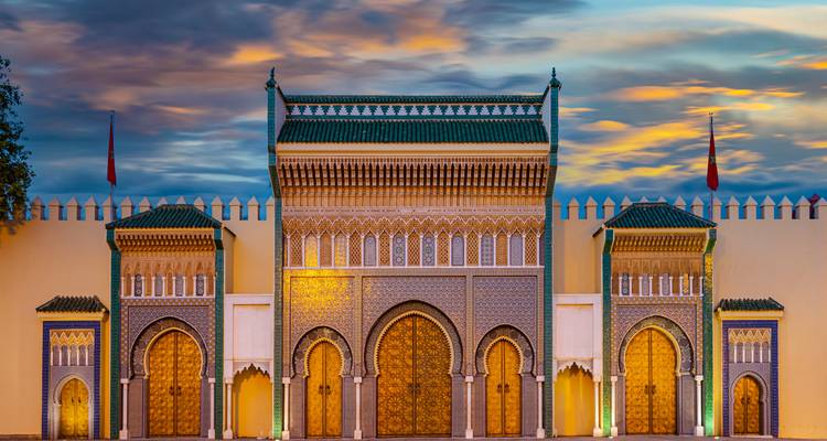 Puertas de mosaico iluminadas del Palacio Real en Fez resplandeciendo bajo un cielo dramático de atardecer.