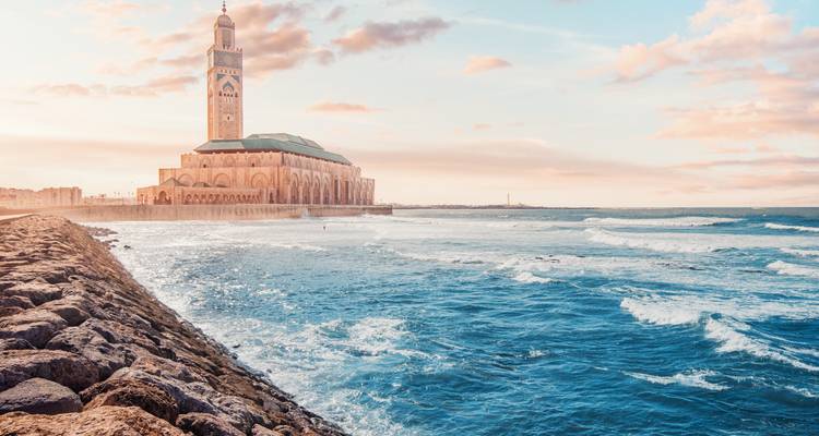 La Mezquita Hassan II se alza junto a las olas rompientes del Atlántico bajo nubes de atardecer en tonos pastel.