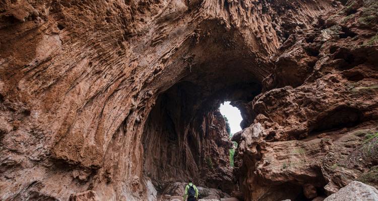 Un excursionista se encuentra debajo de un vasto arco de roca natural dentro de una cueva rugosa de piedra roja.